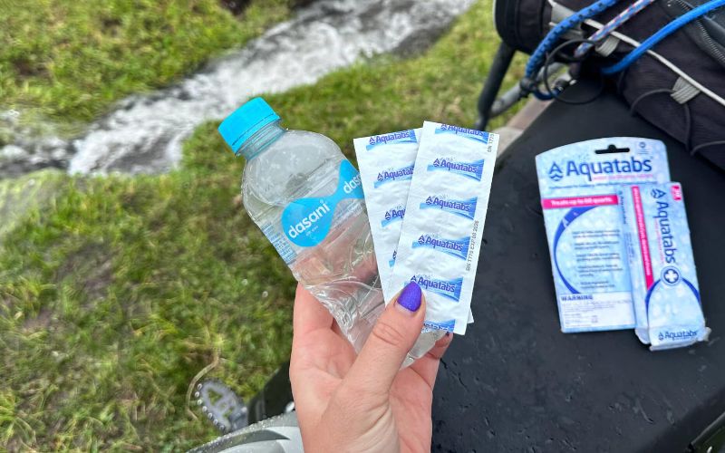 A hand holding water purification tablets and a water bottle outdoors. 